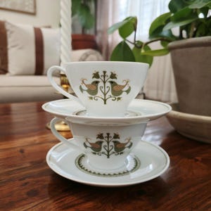May include: Two white teacups and saucers stacked on a wooden table. Each cup and saucer features a green and brown bird and floral design. The cups have a curved handle and a wide rim.