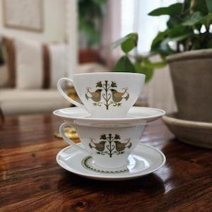 May include: Two white teacups and saucers stacked on a dark wooden table. Each cup and saucer features a decorative design of two birds and foliage in green and gold. The teacups have curved handles.