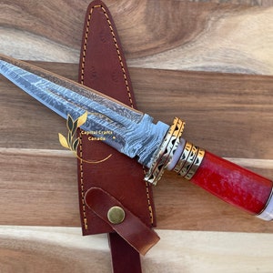 May include: A red and white handled dagger with a gold-colored metal guard and a damascus steel blade. The dagger is in a brown leather sheath.