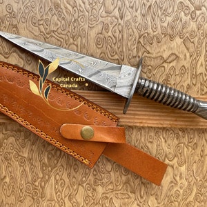 May include: A silver dagger with a patterned blade and a brown leather sheath. The dagger has a twisted handle and a pointed tip. The sheath has a decorative design and a button closure. The dagger and sheath are on a wooden surface.