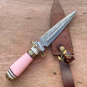 May include: A Damascus steel dagger with a pink handle and a brown leather sheath. The blade has a unique pattern, and the handle is accented with gold and white details. The sheath has a strap with a gold emblem that says "Capital Crafts Canada."