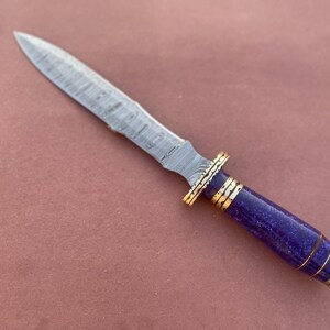 May include: A Damascus steel dagger with a purple and gold handle. The blade has a wavy pattern and the handle is made of a purple material with gold accents.