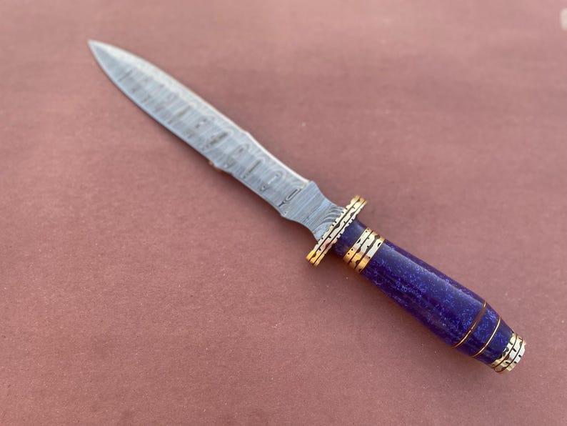 May include: A Damascus steel dagger with a purple and gold handle. The blade has a wavy pattern and the handle is made of a purple material with gold accents.