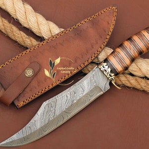 May include: A Damascus steel hunting knife with a brown wood handle and a leather sheath. The knife has a decorative brass guard and pommel. The sheath has the text "Capital Crafts Canada" embossed on it.
