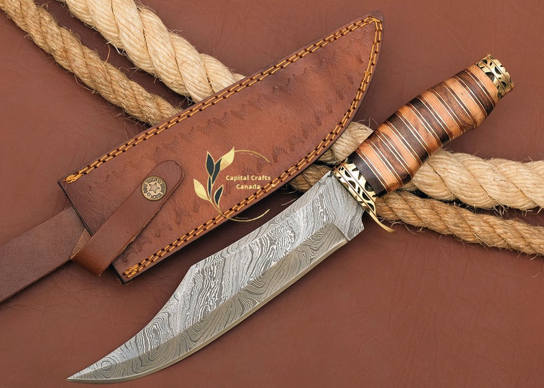 May include: A Damascus steel hunting knife with a brown wood handle and a leather sheath. The knife has a decorative brass guard and pommel. The sheath has the text "Capital Crafts Canada" embossed on it.