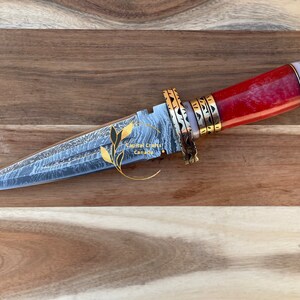 Op de afbeelding: Een decoratieve dolk met een stalen lemmet met patroon en een rode, witte en gouden handgreep. De handgreep heeft een rood steenachtig materiaal, witte delen en goudkleurige metalen accenten. Het lemmet heeft een uniek patroon. De tekst "Capital Crafts Canada" is zichtbaar.