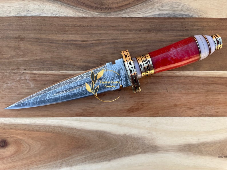 Op de afbeelding: Een decoratieve dolk met een stalen lemmet met patroon en een rode, witte en gouden handgreep. De handgreep heeft een rood steenachtig materiaal, witte delen en goudkleurige metalen accenten. Het lemmet heeft een uniek patroon. De tekst "Capital Crafts Canada" is zichtbaar.