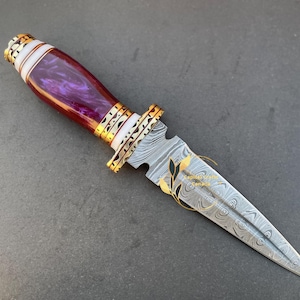 May include: A purple and white handle dagger with a Damascus steel blade. The blade has a decorative pattern and is sharpened to a point. The handle is wrapped in white and purple resin and has gold accents.