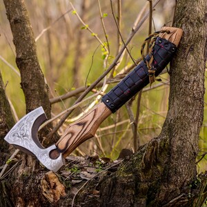 May include: A hand-forged axe with a wooden handle and a leather wrap. The axe head has a detailed design and is resting on a tree stump in a forest setting.