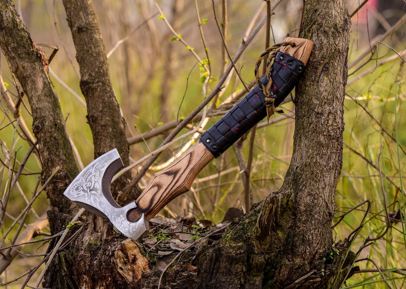 May include: A hand-forged axe with a wooden handle and a leather wrap. The axe head has a detailed design and is resting on a tree stump in a forest setting.