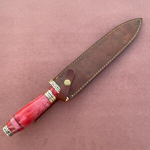 May include: A knife with a red and white handle and a brown leather sheath. The sheath has a brass button closure.