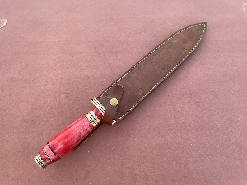 May include: A knife with a red and white handle and a brown leather sheath. The sheath has a brass button closure.