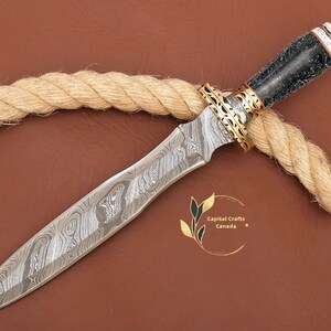 May include: A Damascus steel dagger with a black and white handle and gold accents. The dagger is lying on a brown surface with a rope.