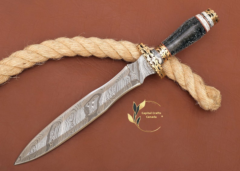 May include: A Damascus steel dagger with a black and white handle and gold accents. The dagger is lying on a brown surface with a rope.