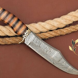 May include: A Damascus steel hunting knife with a wooden handle featuring a striped pattern. The handle is decorated with gold accents. The knife is lying on a brown surface with a rope.
