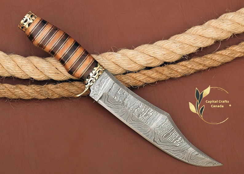 May include: A Damascus steel hunting knife with a wooden handle featuring a striped pattern. The handle is decorated with gold accents. The knife is lying on a brown surface with a rope.