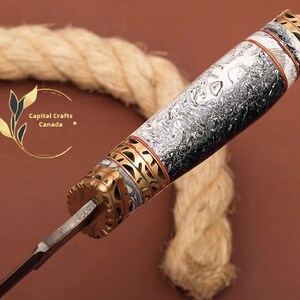 May include: Close-up of a knife handle with a black and white marbled pattern, gold accents, and a red and white band. The handle is made of wood and has a decorative design. Capital Crafts Canada is printed on the image.