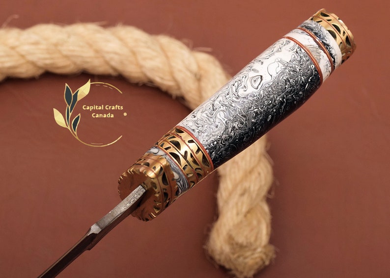 May include: Close-up of a knife handle with a black and white marbled pattern, gold accents, and a red and white band. The handle is made of wood and has a decorative design. Capital Crafts Canada is printed on the image.