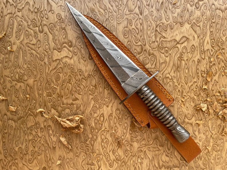 Op de afbeelding: Een Damascus stalen dolk met een puntig lemmet en een spiraalvormige handgreep. De dolk ligt op een bruine leren schede, tegen een achtergrond van houtnerf. Het lemmet heeft een opvallend patroon.