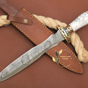 May include: A white and brown patterned dagger with a gold-coloured handle and a leather sheath. The dagger has a Damascus steel blade and is wrapped in a rope.