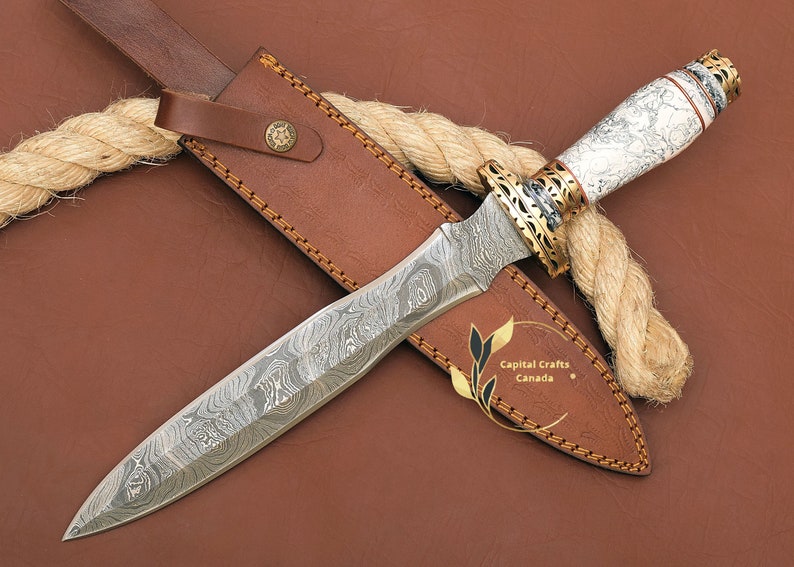 May include: A white and brown patterned dagger with a gold-coloured handle and a leather sheath. The dagger has a Damascus steel blade and is wrapped in a rope.