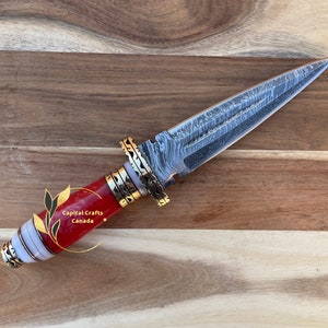 Op de afbeelding: Een decoratieve dolk met een stalen lemmet met patroon en een handvat met rode, witte en gouden accenten. Het handvat is gemaakt van rood materiaal met witte en gouden banden. Het lemmet heeft een uniek patroon. De dolk wordt tentoongesteld op een houten oppervlak.
