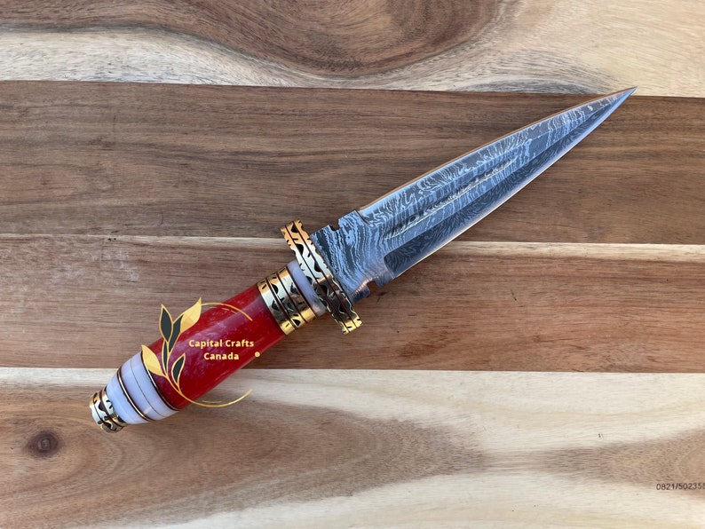 Op de afbeelding: Een decoratieve dolk met een stalen lemmet met patroon en een handvat met rode, witte en gouden accenten. Het handvat is gemaakt van rood materiaal met witte en gouden banden. Het lemmet heeft een uniek patroon. De dolk wordt tentoongesteld op een houten oppervlak.