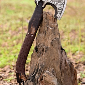 May include: A silver axe with a leather-wrapped wooden handle and a chain. The axe head has intricate carvings and a symbol of a triangle with a circle inside. The axe is resting on a weathered tree stump.