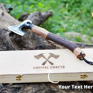 May include: A hand-forged Viking axe with a leather wrapped handle and a wooden box with the logo "Capital Crafts" engraved on the lid. The axe is resting on a wooden log.
