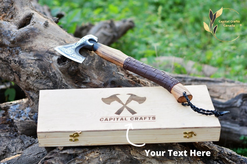 May include: A hand-forged Viking axe with a leather wrapped handle and a wooden box with the logo "Capital Crafts" engraved on the lid. The axe is resting on a wooden log.