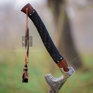 May include: A silver axe with a leather wrapped handle and a tassel. The axe has intricate carvings and a leather strap. The axe is resting on a wooden stump.