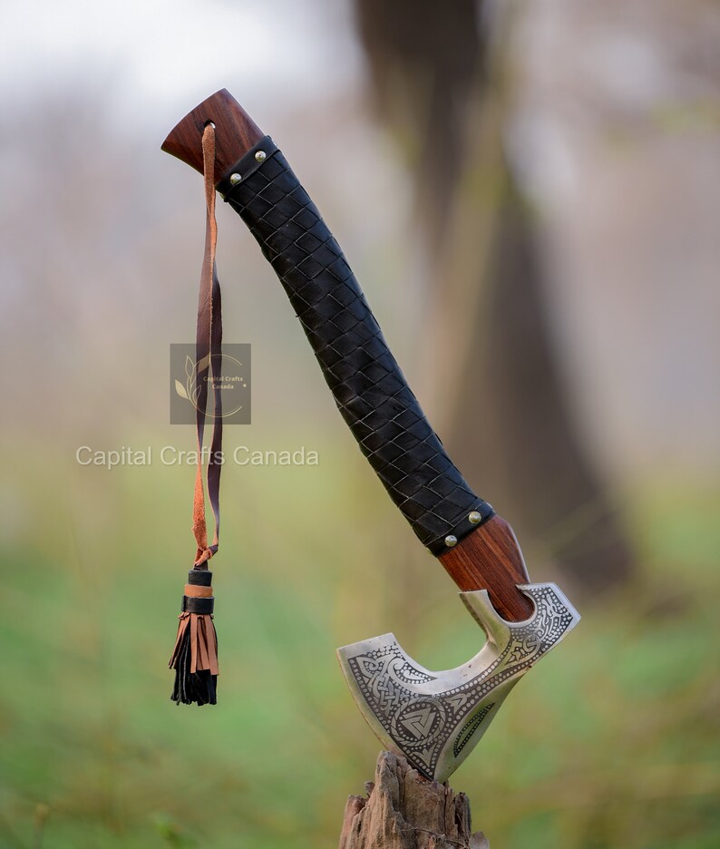 May include: A silver axe with a leather wrapped handle and a tassel. The axe has intricate carvings and a leather strap. The axe is resting on a wooden stump.