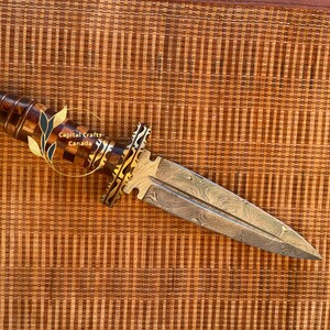Op de afbeelding: Een decoratieve dolk met een patroon op het lemmet en een handvat van hout en goudkleurig metaal. Het handvat heeft een dambordpatroon. Het lemmet heeft een uniek patroon. De dolk wordt getoond op een geweven oppervlak. De tekst "Capital Crafts Canada" is zichtbaar.