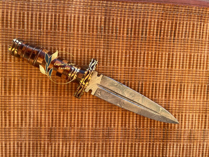 Op de afbeelding: Een decoratieve dolk met een patroon op het lemmet en een handvat van hout en goudkleurig metaal. Het handvat heeft een dambordpatroon. Het lemmet heeft een uniek patroon. De dolk wordt getoond op een geweven oppervlak. De tekst "Capital Crafts Canada" is zichtbaar.