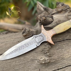 May include: An intricately designed dagger with a silver blade and a handle featuring a cream-coloured grip and copper-coloured accents. The blade is etched with a floral pattern, and the handle has detailed engravings. The dagger rests on weathered wood.