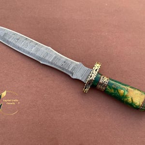 May include: A silver dagger with a green and gold handle. The blade is decorated with a Damascus pattern. The handle is made of wood and has a brass accent.