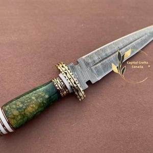 May include: A handcrafted dagger with a patterned steel blade and a green and white handle. The handle is accented with gold-colored metal rings. The blade has a pointed tip and a decorative pattern. The dagger is a collectible item.