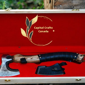 May include: A hand-forged axe with a wooden handle and a leather wrap. The axe is in a wooden box with a red velvet lining. The box has a gold logo that says "Capital Crafts Canada".