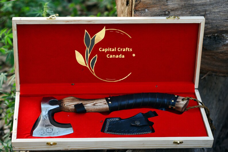 May include: A hand-forged axe with a wooden handle and a leather wrap. The axe is in a wooden box with a red velvet lining. The box has a gold logo that says "Capital Crafts Canada".
