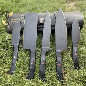 May include: A set of five black knives with dark handles and decorative circular inlays, arranged in front of a black leather roll. The text Capital Crafts Canada is visible on the left side. The knives feature varied blade shapes including a cleaver.