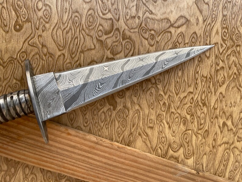 Op de afbeelding: Een dolk van Damascusstaal met een patroon op het lemmet en een zilverkleurige pareerstang. De handgreep is omwikkeld met een getextureerd, zilverkleurig materiaal. Het lemmet heeft een scherpe punt en een kenmerkend wervelpatroon. De dolk wordt gepresenteerd tegen een achtergrond van houtnerf.