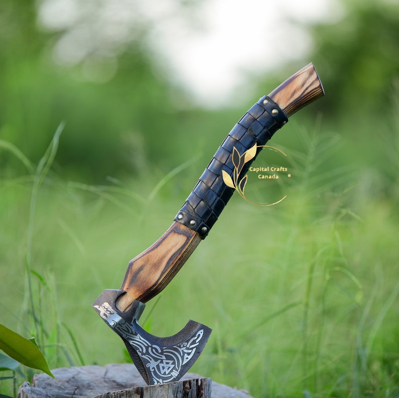 May include: A hand-forged axe with a wooden handle wrapped in black leather. The axe head features a decorative design and the handle has a brass rivet. The axe is resting on a wooden stump in a grassy field.