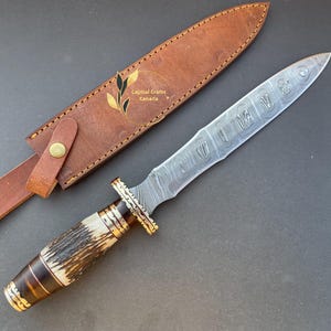 May include: A Damascus steel dagger with a brown and white stag horn handle and a leather sheath. The blade has a decorative pattern and the sheath has the text "Capital Crafts Canada".