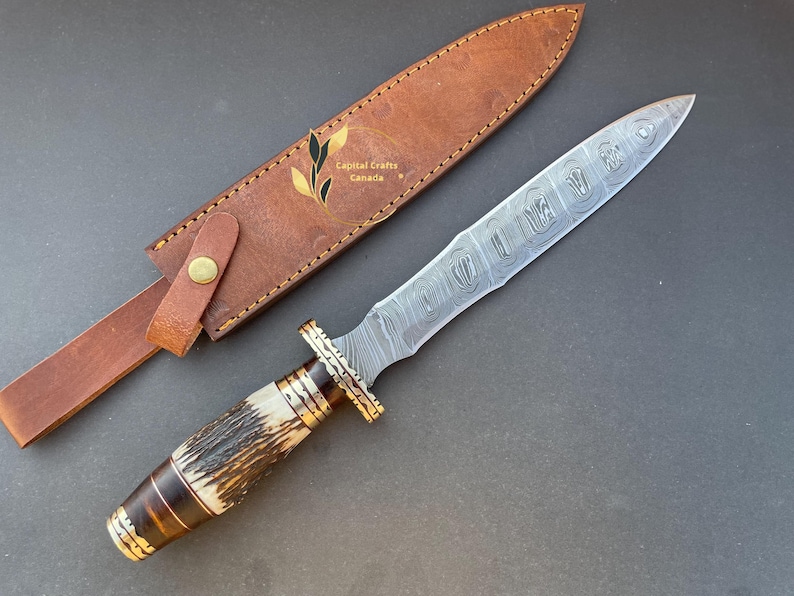 May include: A Damascus steel dagger with a brown and white stag horn handle and a leather sheath. The blade has a decorative pattern and the sheath has the text "Capital Crafts Canada".