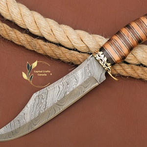Può includere: Un coltello da caccia in acciaio damasco con manico in legno e ottone. Il coltello è appoggiato su una superficie marrone con una corda.