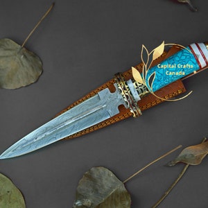 Op de afbeelding: Een handgemaakte dolk met een patroon op het lemmet en een turquoise en witte handgreep. De dolk zit in een bruine leren schede. De handgreep heeft goudkleurige accenten. De tekst "Capital Crafts Canada" is zichtbaar.