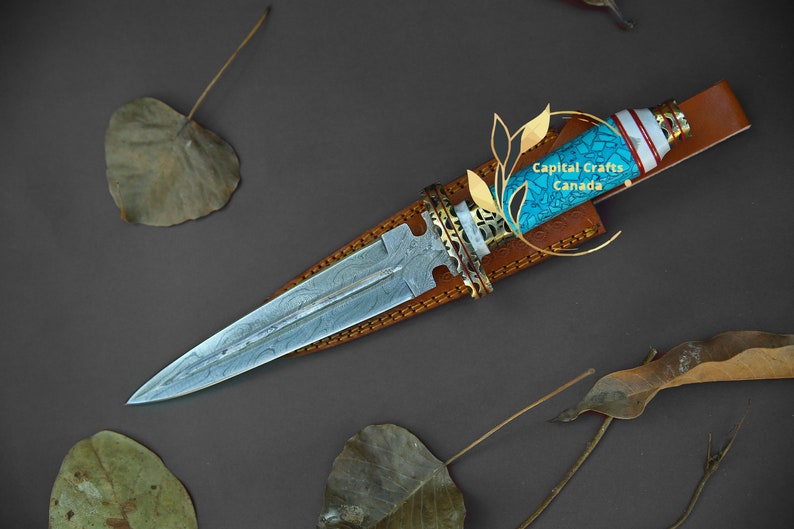 Op de afbeelding: Een handgemaakte dolk met een patroon op het lemmet en een turquoise en witte handgreep. De dolk zit in een bruine leren schede. De handgreep heeft goudkleurige accenten. De tekst "Capital Crafts Canada" is zichtbaar.