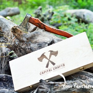May include: A wooden box with a gold engraved axe design and the text "CAPITAL CRAFTS". The box is open and a small axe with a wooden handle is resting on top of the box. The axe has a gold eagle design on the blade.