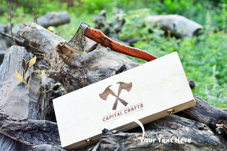 May include: A wooden box with a gold engraved axe design and the text "CAPITAL CRAFTS". The box is open and a small axe with a wooden handle is resting on top of the box. The axe has a gold eagle design on the blade.