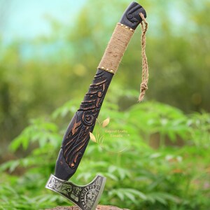 May include: A black wooden axe with a carved handle and a silver blade. The blade has a skull and runes engraved on it. The axe is on a wooden stump with a green background.